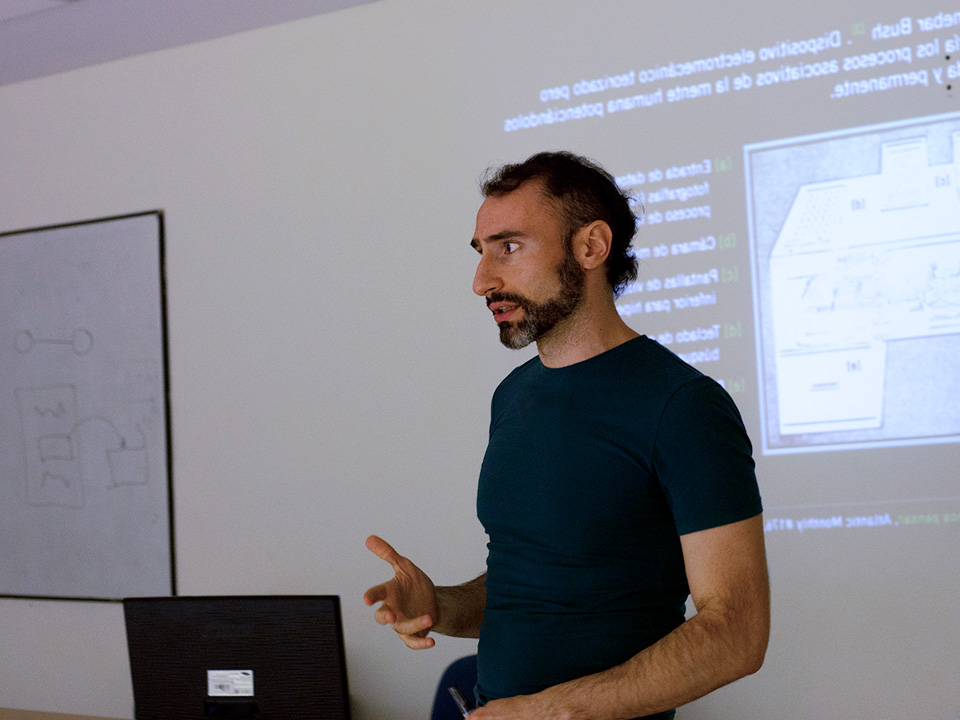 Profesor con barba y remera verde al frente de una clase, se ve una diapositiva proyectada en el fondo